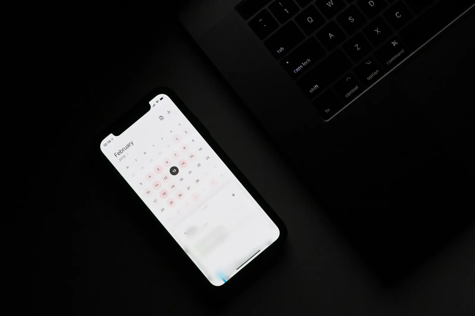 A smartphone showing a calendar app, lying on a black desk beside a laptop keyboard.